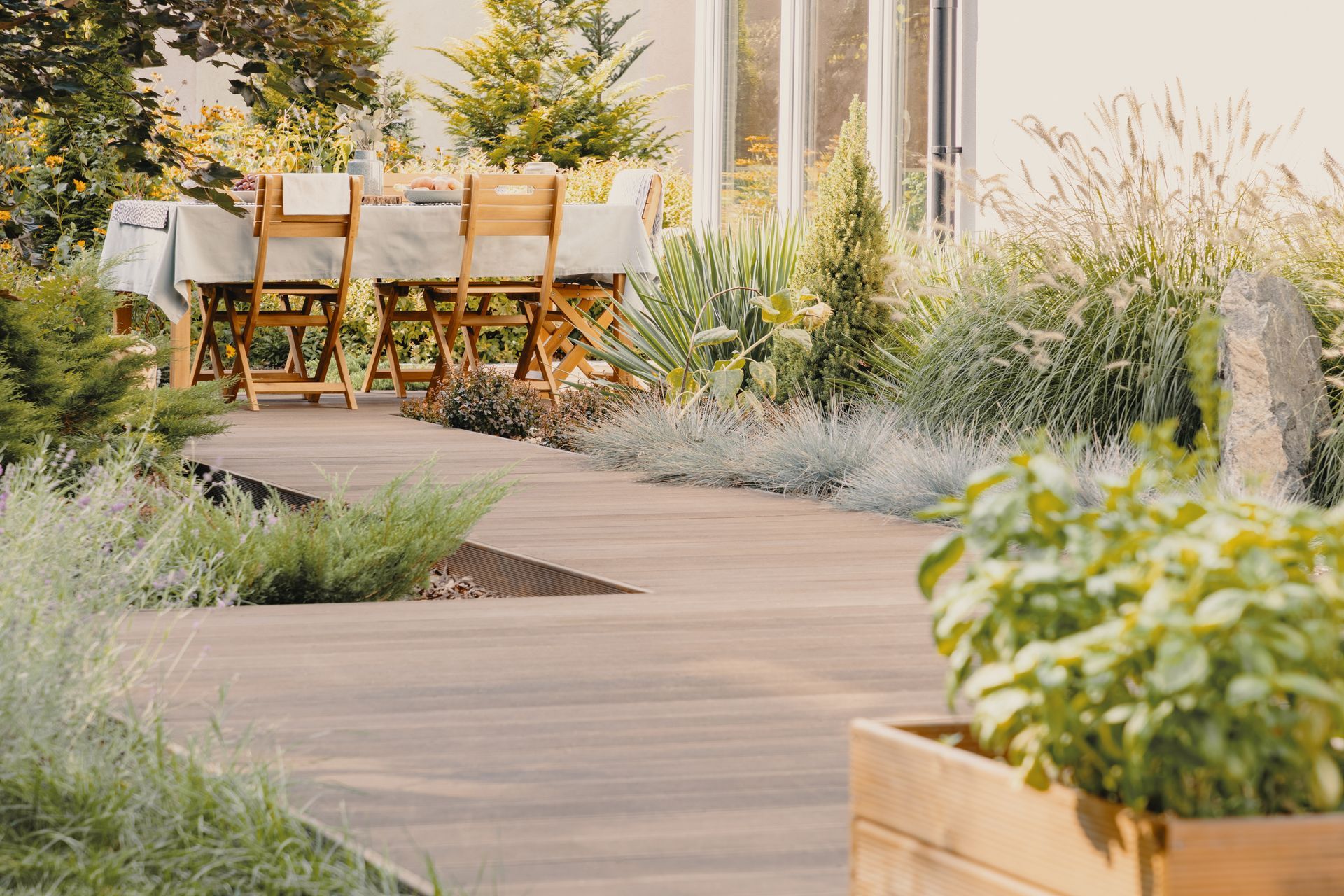 Une terrasse en bois mène à une table à manger au milieu de la végétation, dans un espace extérieur baigné de soleil.