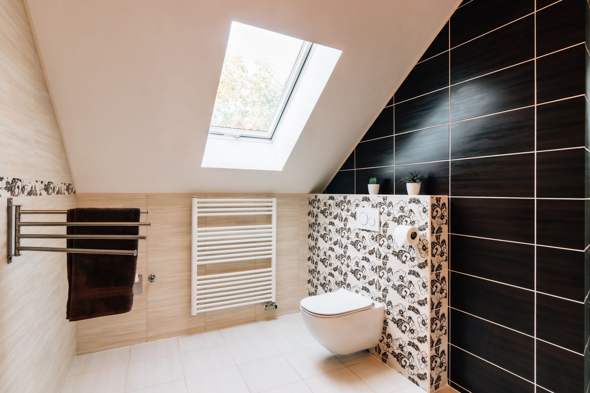 Une salle de bains sous un toit avec un puits de lumière, des toilettes, une crédence à motifs et un porte-serviettes.