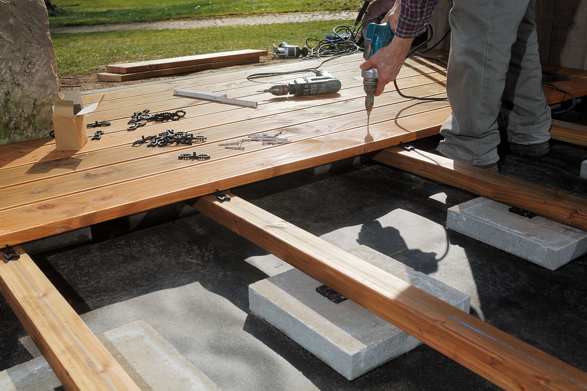 Une personne utilise une perceuse pour installer des lames de terrasse en bois sur un cadre soutenu par des blocs de béton.