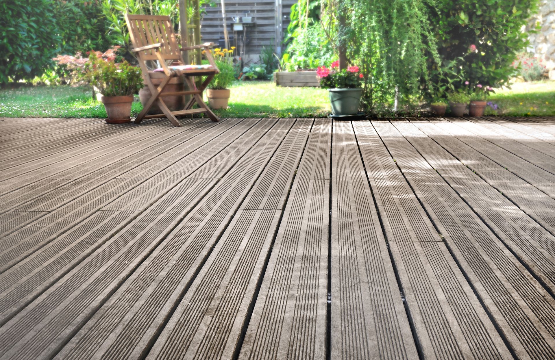 Une terrasse en bois à la surface texturée et rainurée, avec une chaise et plusieurs plantes en pot dans un jardin.