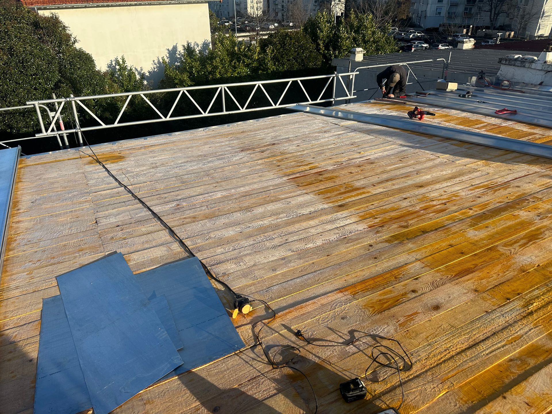 Un ouvrier du bâtiment installe des plaques d'isolation jaunes sur un toit plat, à l'aide d'un échafaudage et d'une bâche.