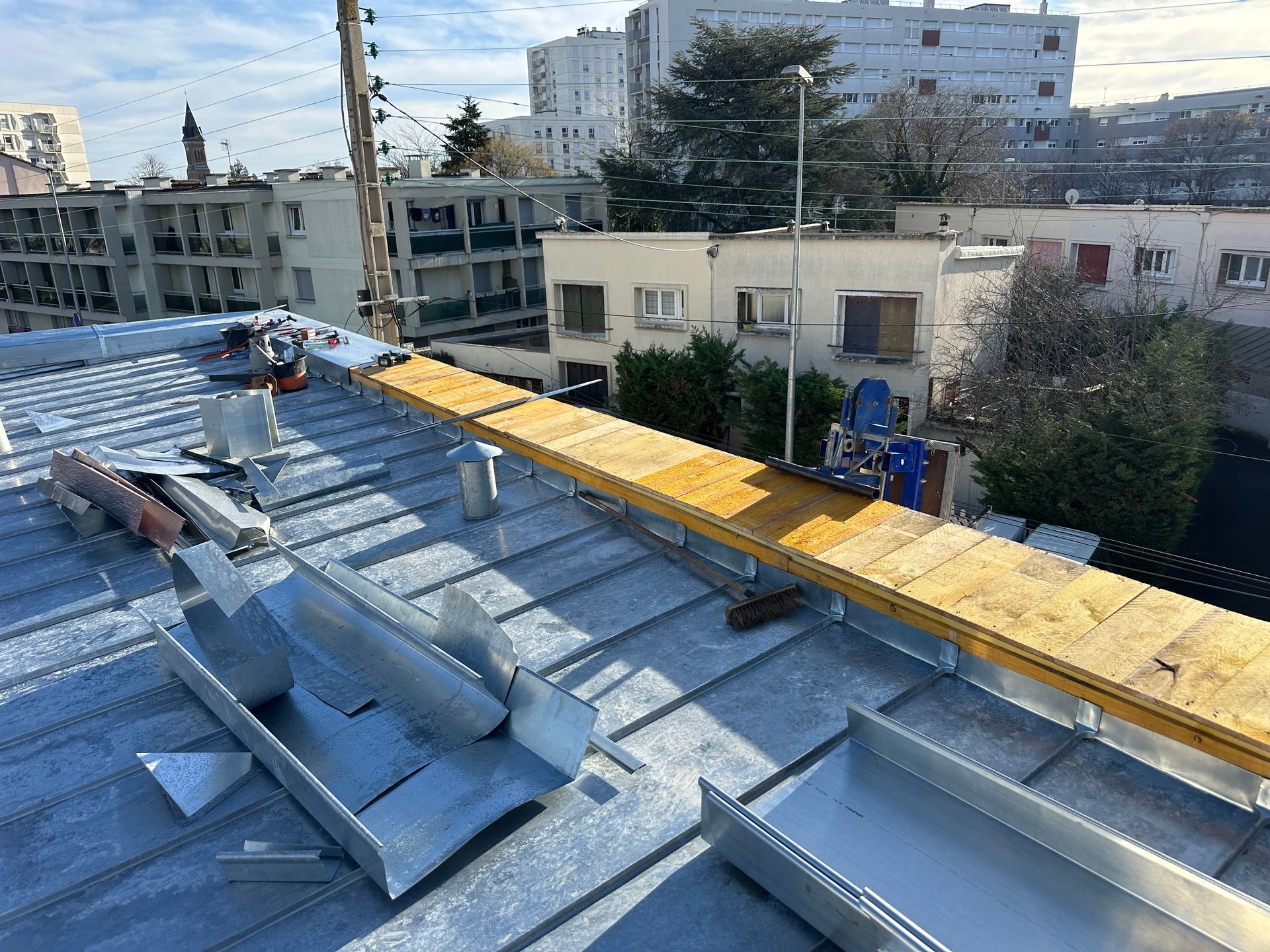 Un toit métallique en construction avec une passerelle en bois installée, se détachant sur un fond urbain de bâtiments.