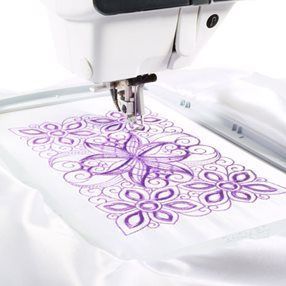 Una máquina de coser está bordando una flor morada sobre una tela blanca.