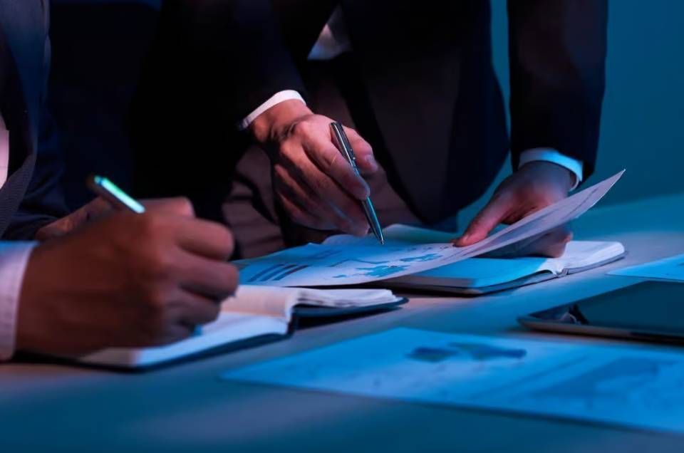 Hände in dunklen Anzügen sichten Dokumente und schreiben auf Papier an einem blau beleuchteten Tisch.