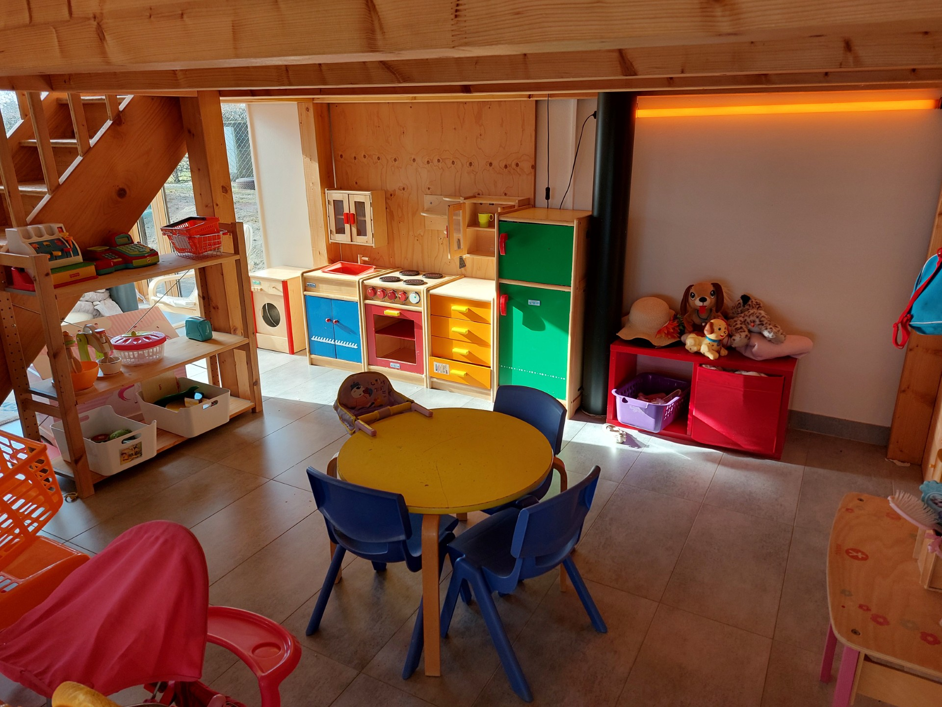 Une salle de jeux avec une cuisine en bois pour enfants, une petite table jaune avec des chaises bleues, un espace de rangement pour les jouets et une mezzanine.