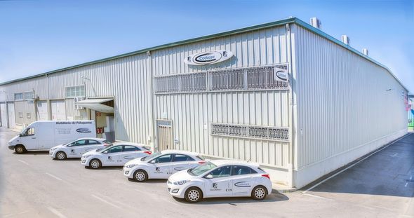 Nave industrial blanca con coches aparcados y una furgoneta de reparto. Los vehículos llevan el logotipo de la empresa en las puertas.