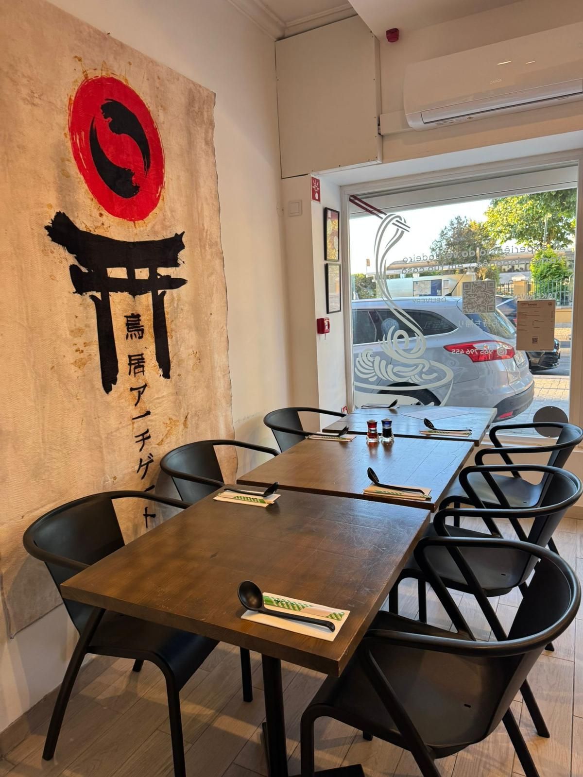 Interior de restaurante japonês com mesas, cadeiras pretas e uma janela com vista para a rua. Uma faixa pendurada na parede.