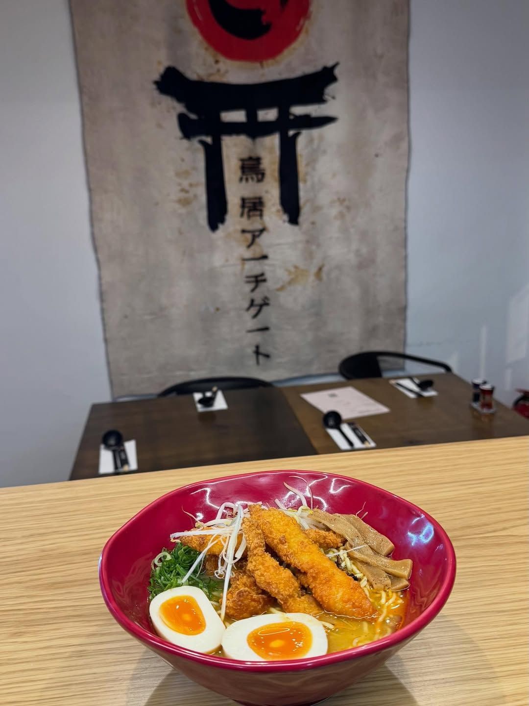 Tigela de ramen com itens fritos, ovo e guarnição verde sobre uma mesa de madeira, com restaurante japonês ao fundo.