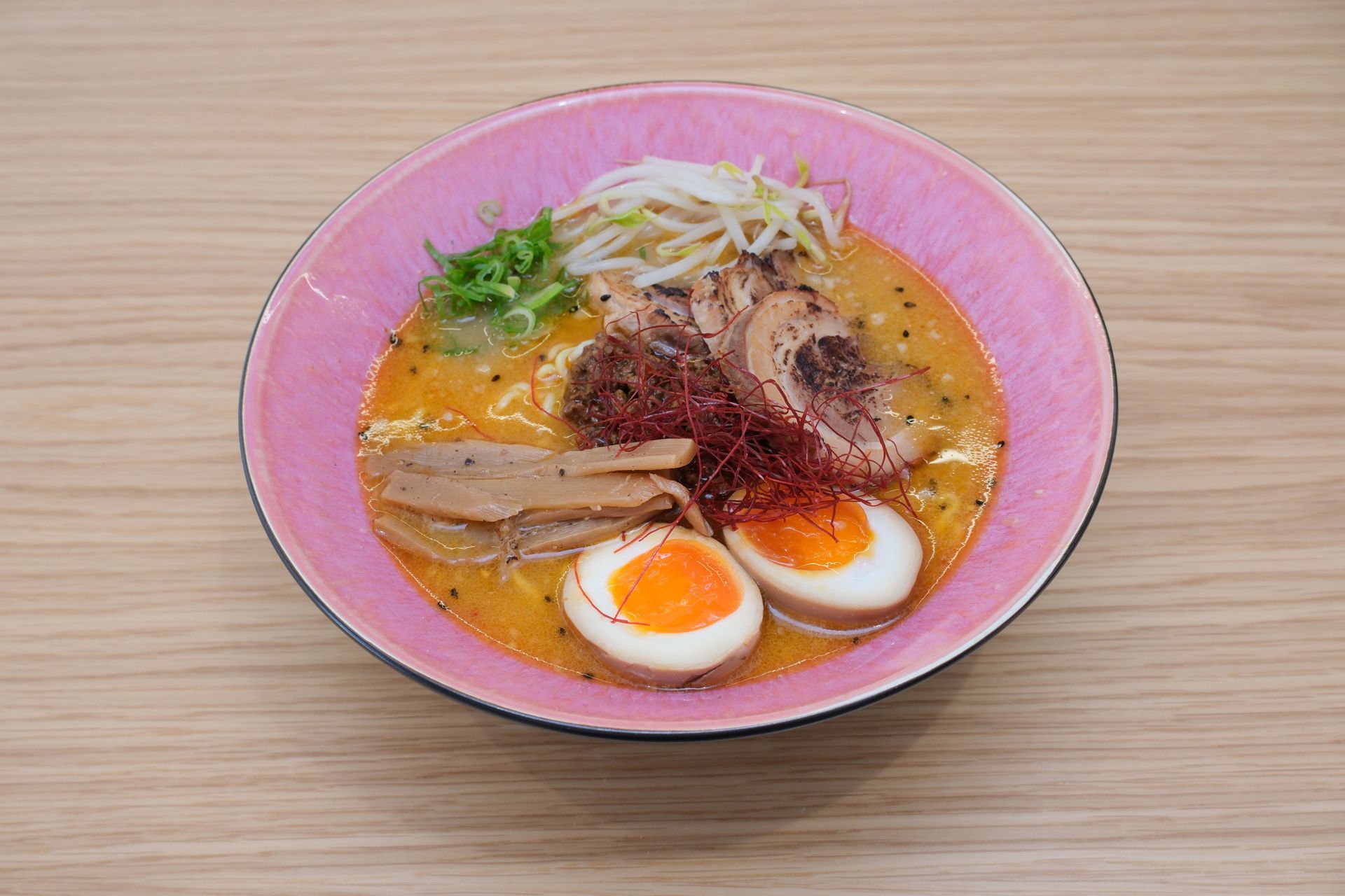 Ramen em uma tigela rosa sobre uma superfície de madeira, com ovo, carne de porco, brotos e molho.