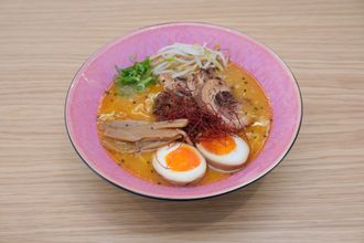 Ramen em uma tigela rosa sobre uma superfície de madeira, com ovo, carne de porco, brotos e molho.