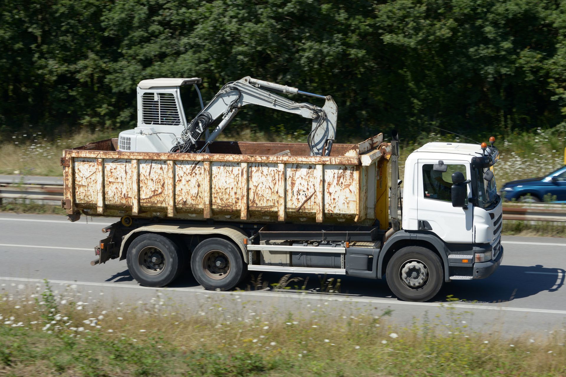 Un camion-benne transportant une excavatrice est stationné en bord de route, devant des arbres.