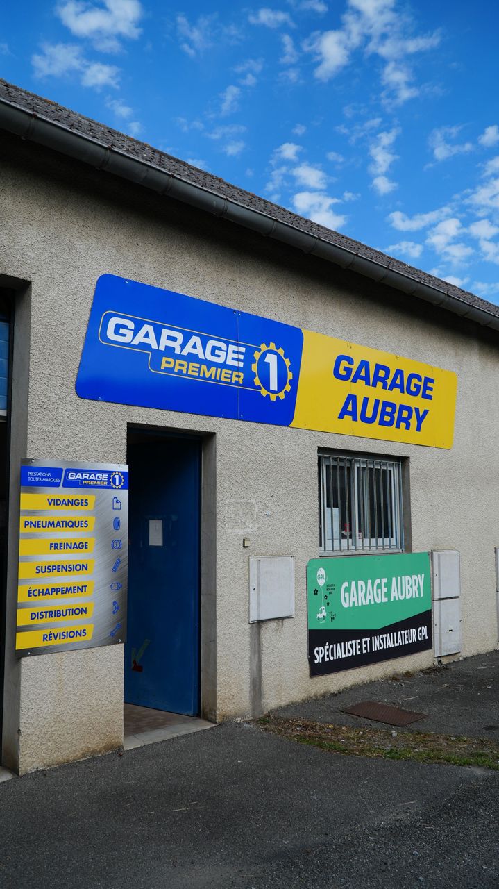 Voitures garées devant un garage, portes de garage ouvertes. Bâtiment beige, parking en gravier, ciel bleu.
