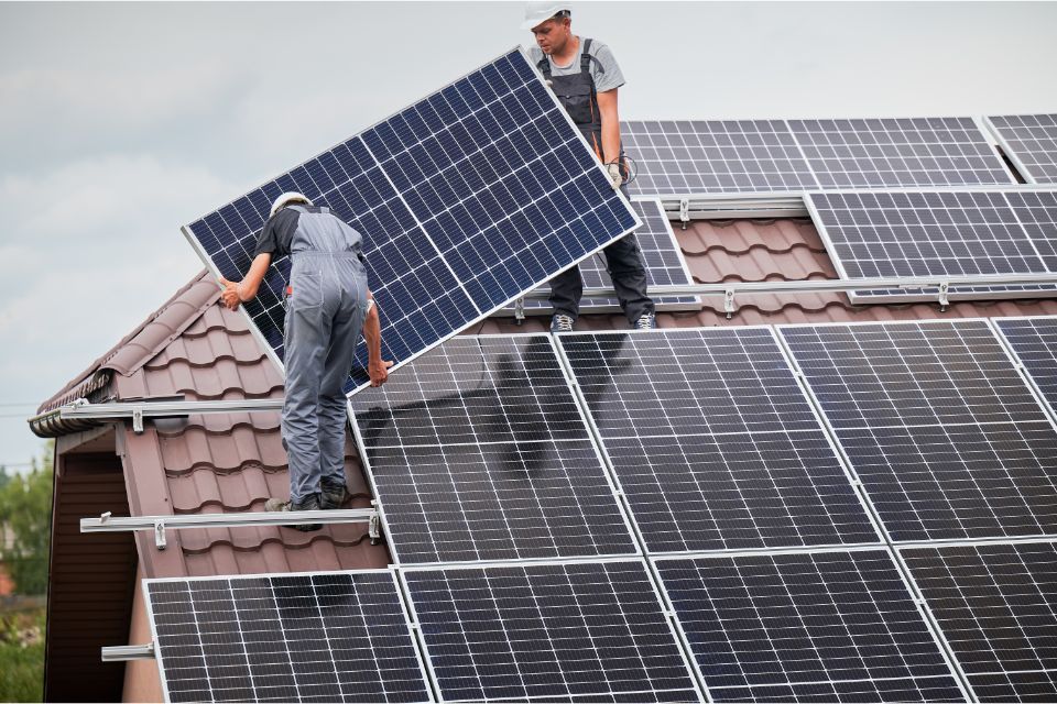 Zwei Mitarbeiter von Saeid Nasouhi heben ein Solarmodul auf ein Dach, auf dem bereits mehrere Anlagen montiert sind