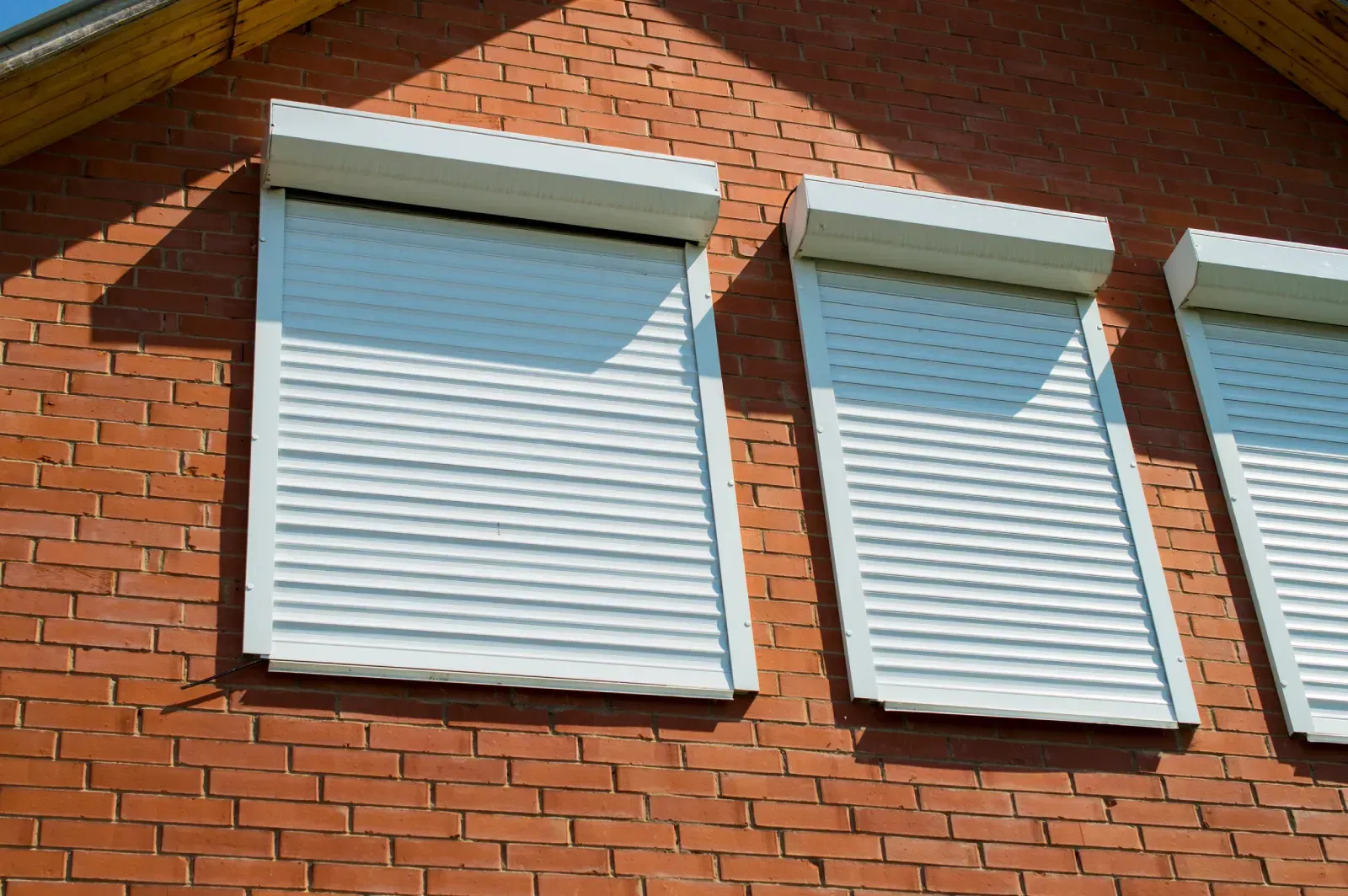 Tres persianas blancas enrollables sobre las ventanas de un edificio de ladrillo rojo, ligeramente inclinadas.