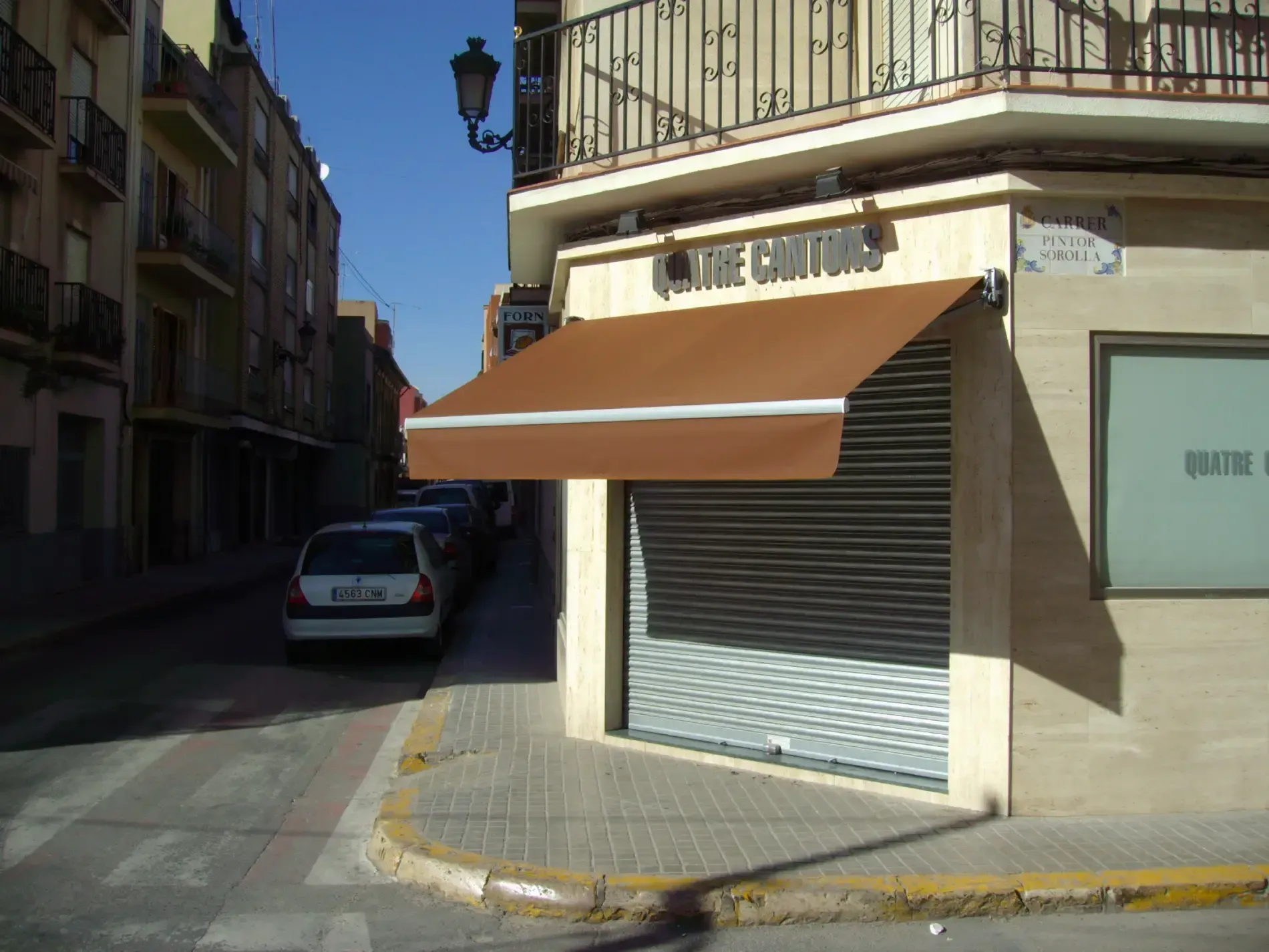 Edificio esquinero con toldo marrón, persiana metálica cerrada, calle con coches aparcados.