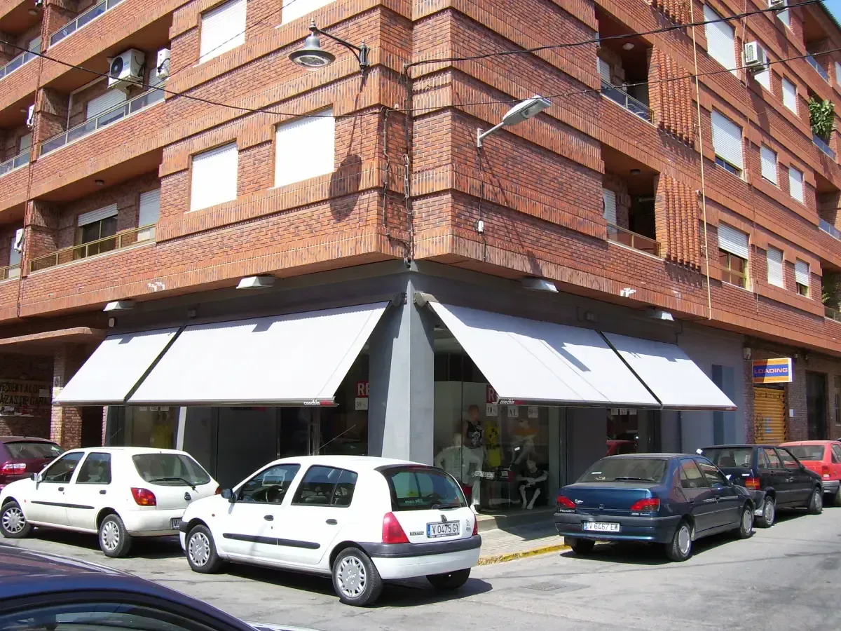 Edificio esquinero con toldos blancos y coches aparcados en la calle. Fachada de ladrillo.