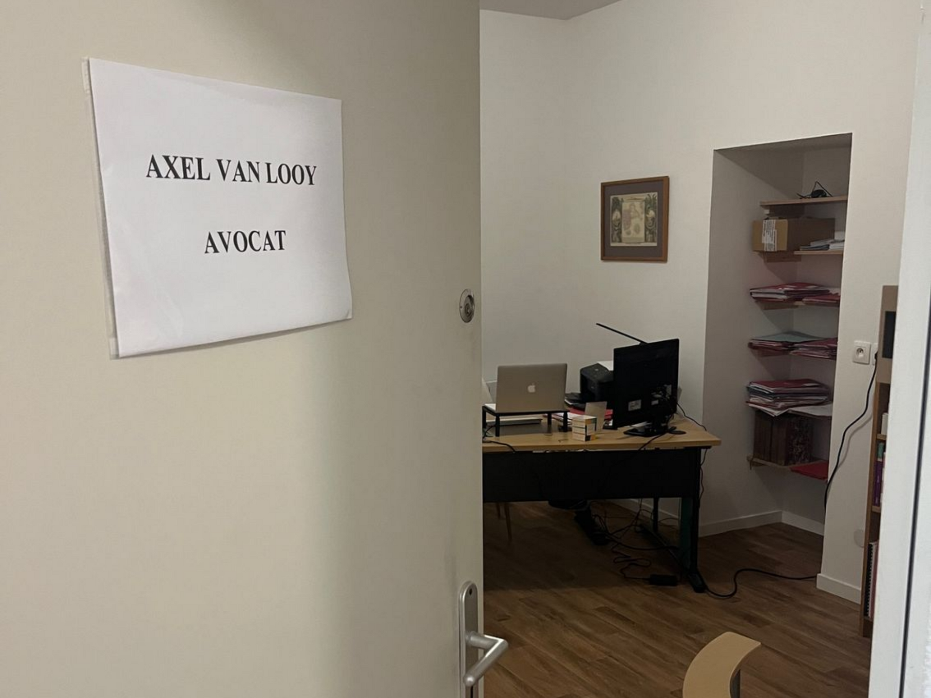 Entrée du bureau avec un panneau indiquant : Axel Van Loov Avocat. Un bureau et des étagères.