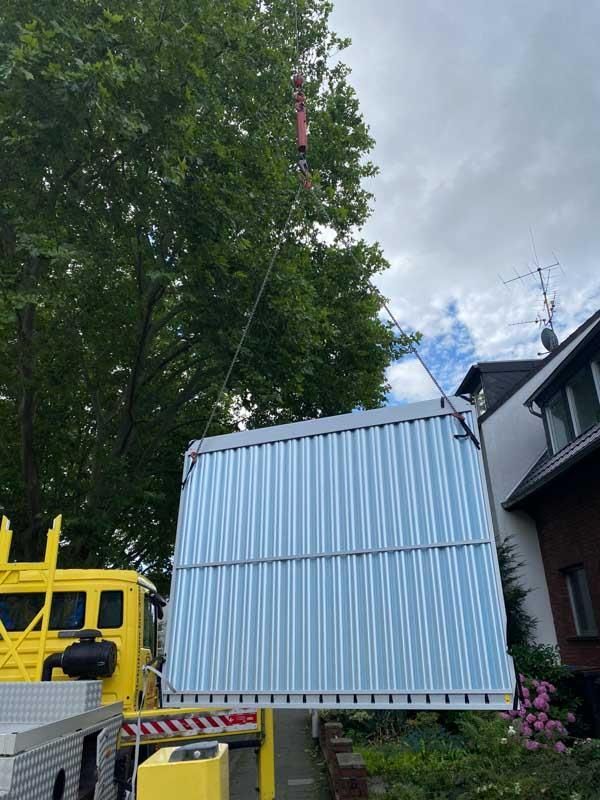 Ein Kran hebt einen blau-weißen Container vor einem Haus an.