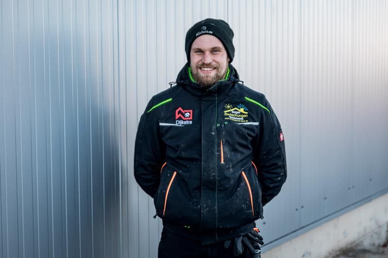 Ein Mann in einer schwarzen Jacke und einem Hut steht vor einer Wand.