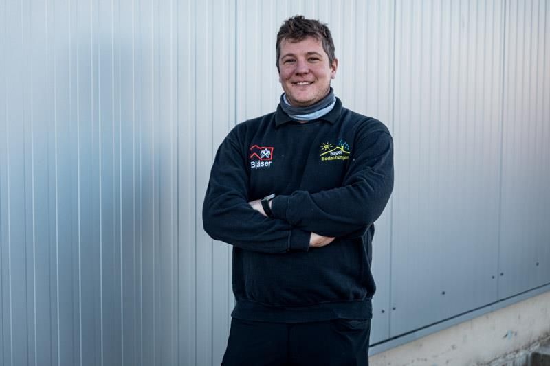 Ein Mann in einem schwarzen Sweatshirt steht mit verschränkten Armen vor einer Wand.