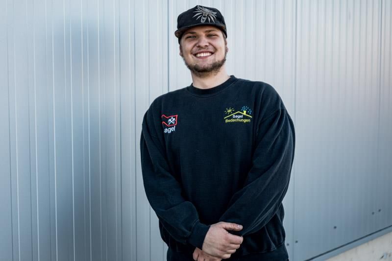 Ein Mann mit einem schwarzen Sweatshirt und einer Mütze steht vor einer Wand.