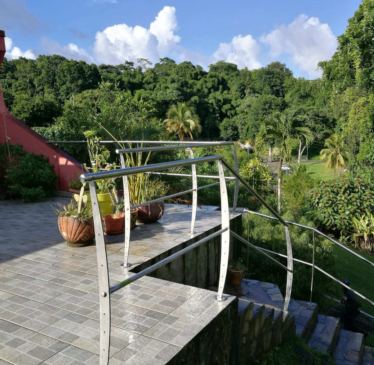 Vue extérieure d'un escalier et d'une allée avec des rambardes métalliques donnant sur une végétation luxuriante par une journée ensoleillée.