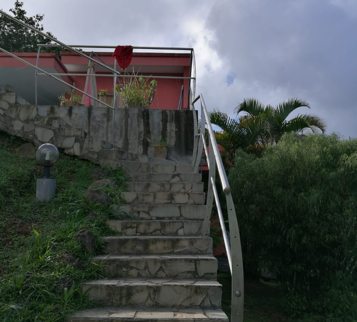 Des marches en pierre mènent à un bâtiment rose doté d'une rambarde en métal, entouré de verdure et d'un ciel nuageux.
