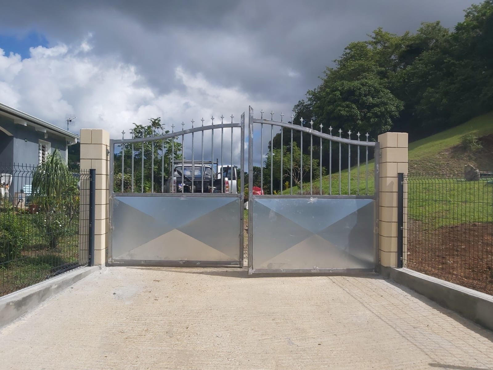 Portail d'allée en métal argenté avec partie supérieure décorative, entre des colonnes de briques sur béton.