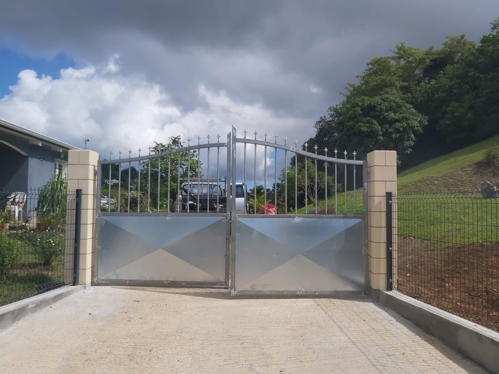 Portail d'allée en métal au design géométrique, placé entre deux piliers couleur crème, sur fond de ciel nuageux.