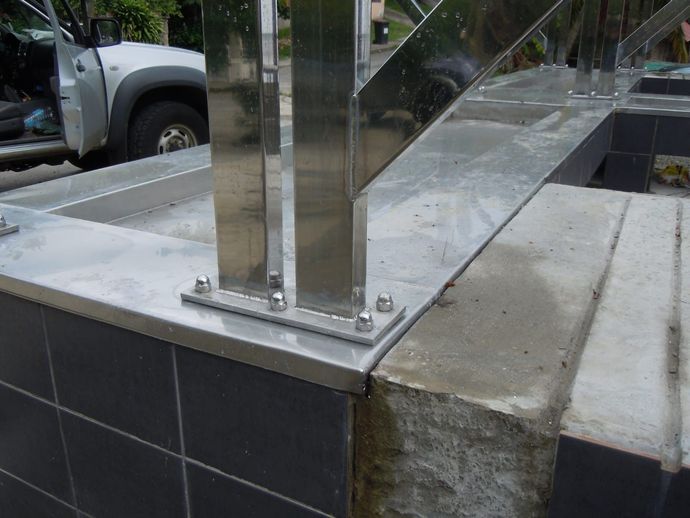 Rambarde en acier inoxydable fixée à un mur carrelé et à une base en béton ; boulons apparents. Un camion est visible en arrière-plan.