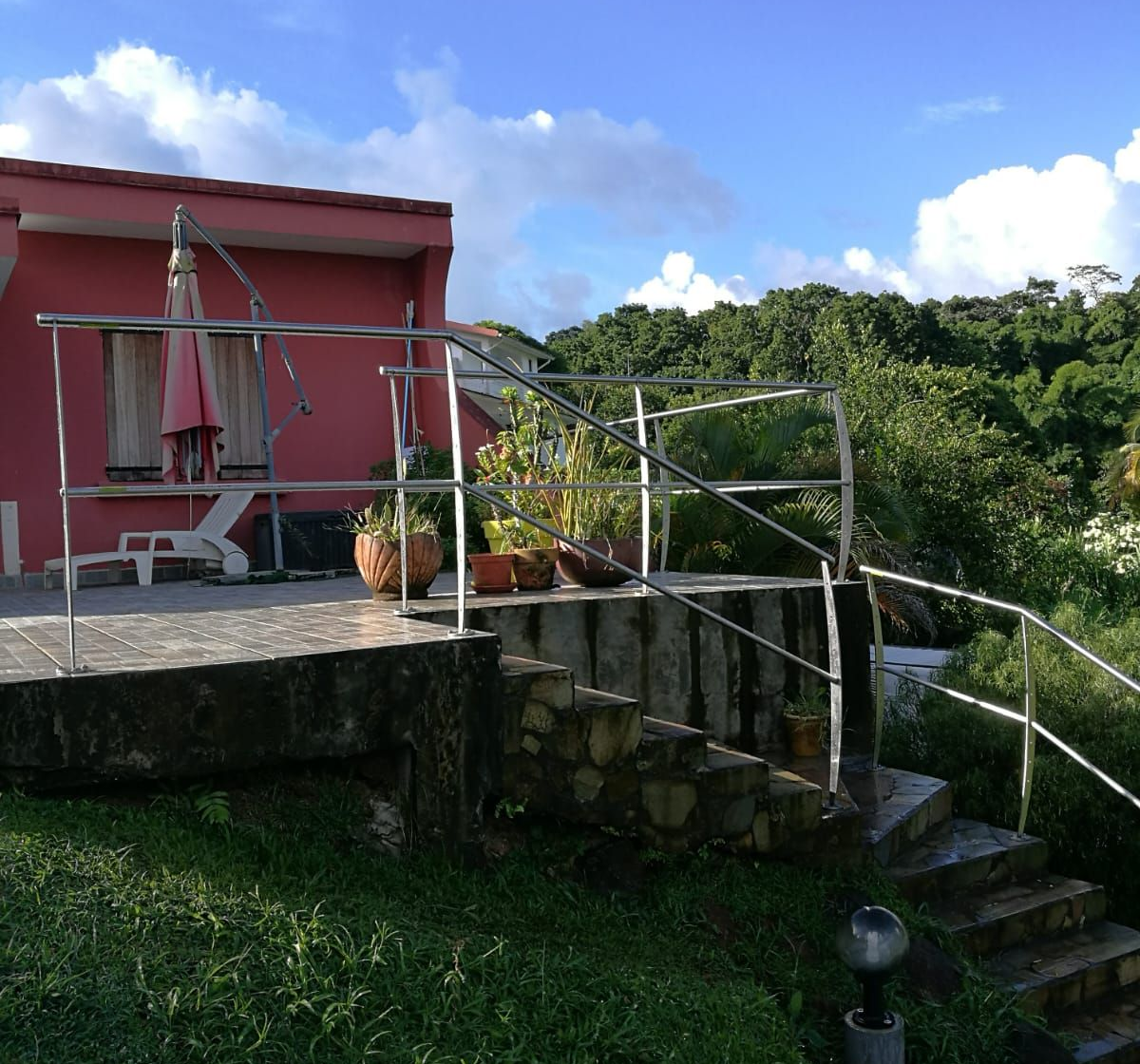 Bâtiment rose avec terrasse, escalier et balustrade en métal donnant sur une végétation luxuriante sous un ciel bleu.