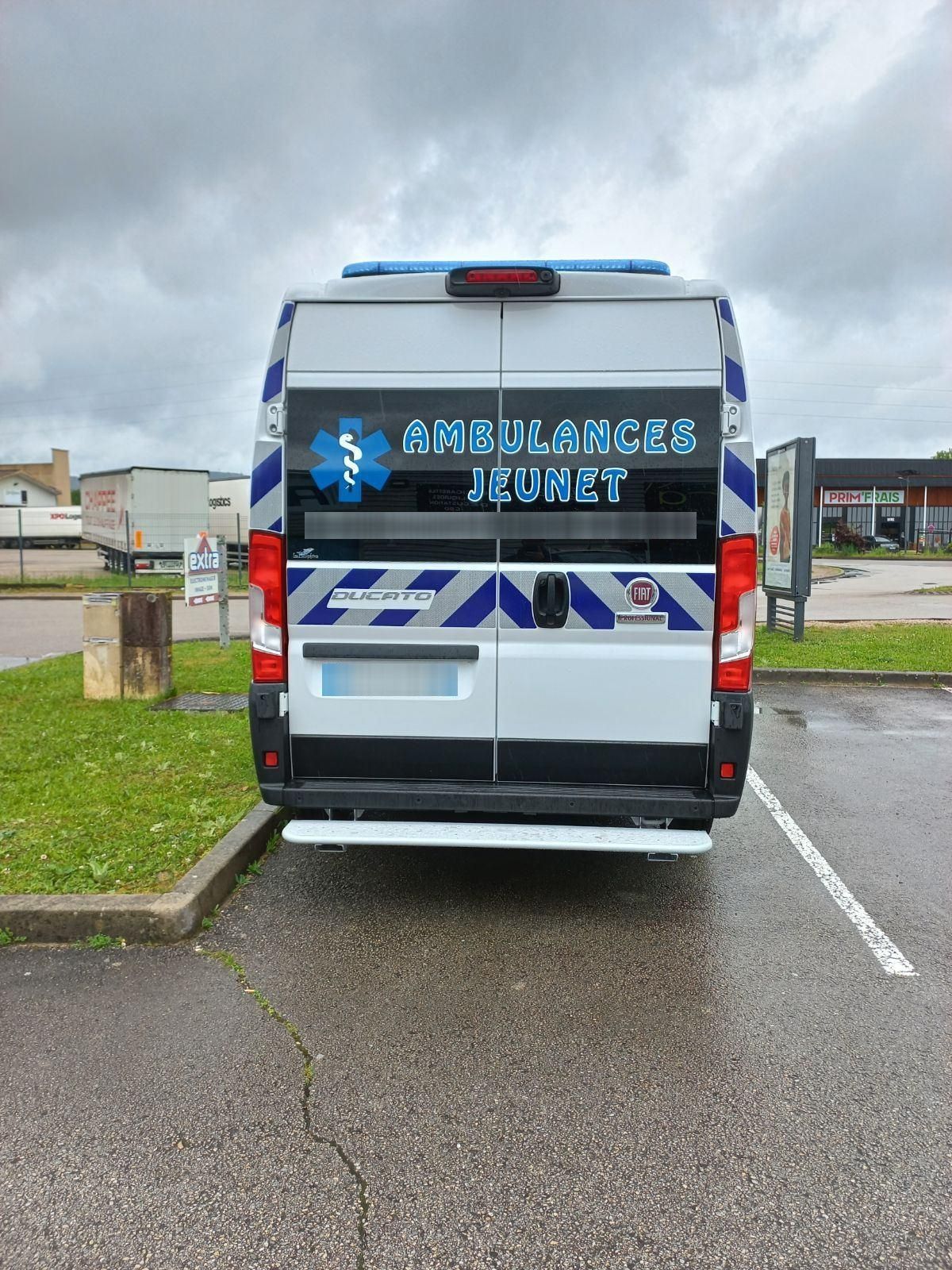 Vue arrière d'une ambulance stationnée sur un parking par temps nuageux. Elle est ornée de rayures et d'inscriptions bleues et blanches.