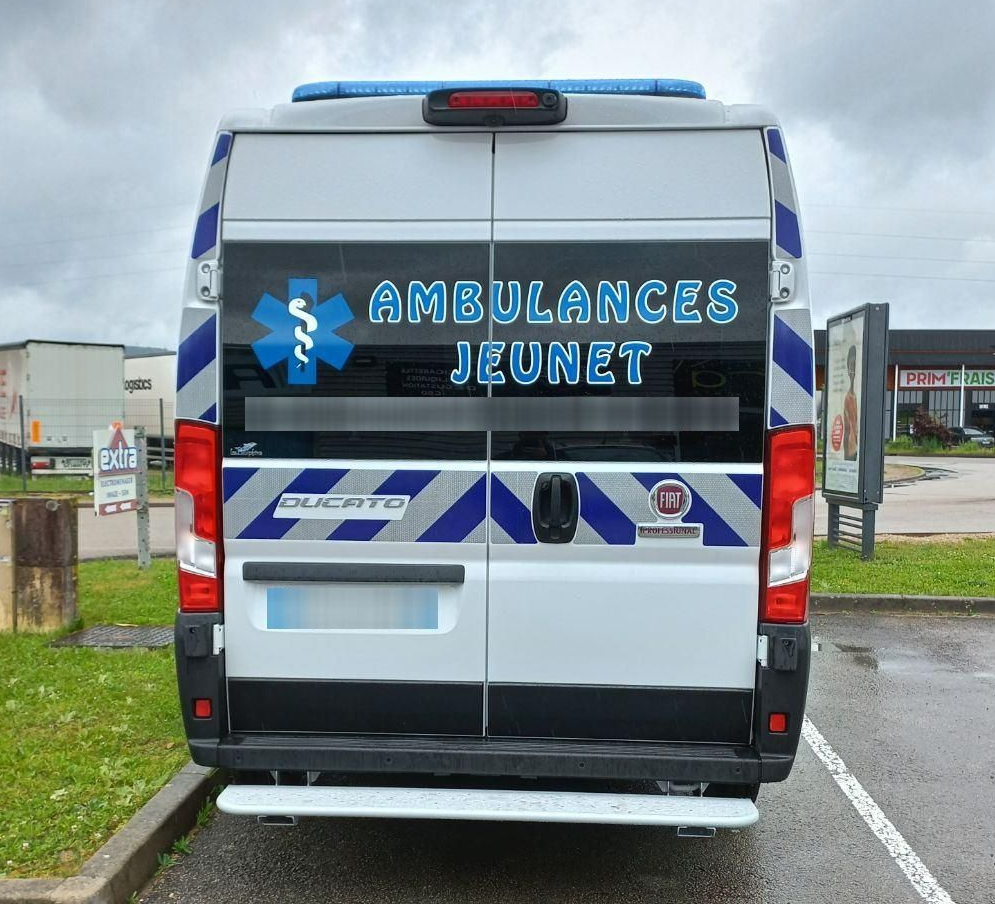Vue arrière d'une ambulance aux marquages ​​bleus et blancs. Inscription : AMBULANCES JEUNET. Temps nuageux.