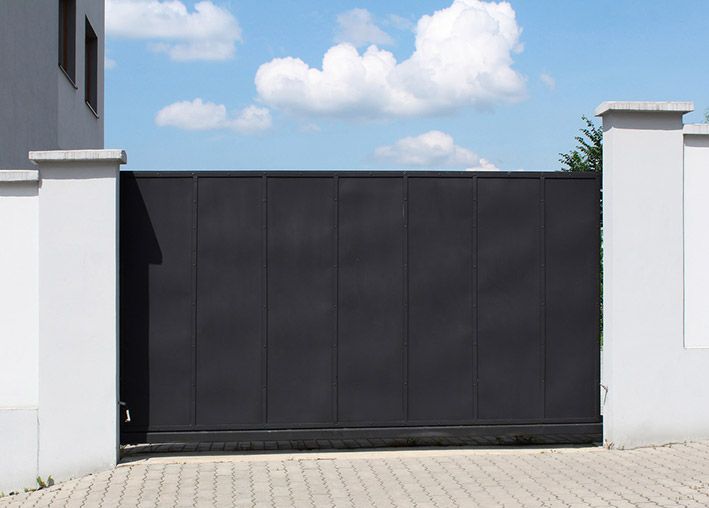 Portail coulissant en métal noir devant un mur blanc et une maison, sous un ciel bleu avec des nuages.