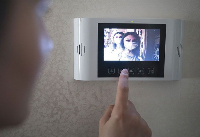 Une personne regarde l'écran d'une sonnette vidéo et appuie sur un bouton. Deux femmes masquées sont visibles à l'écran.