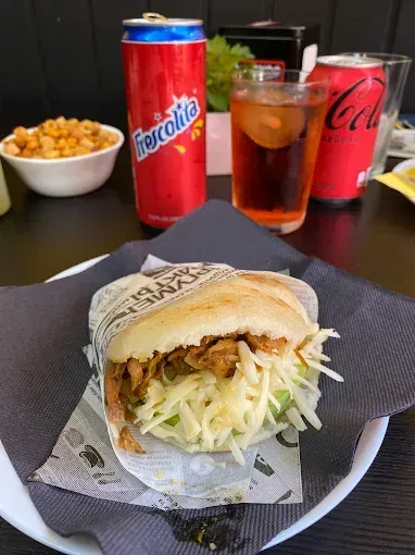 Un sándwich de arepa rellena con queso rallado y carne, con bebidas y bocadillos en una mesa.