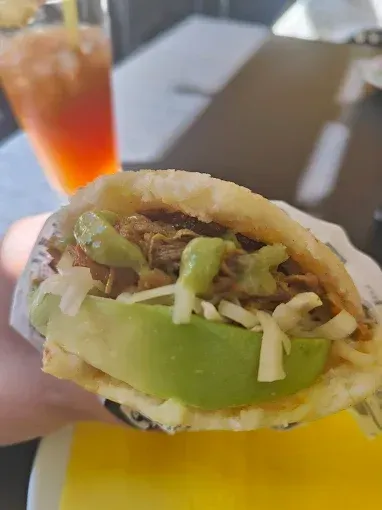 Arepa rellena de aguacate, carne deshebrada y salsa verde, sostenida en la mano; té helado de fondo.