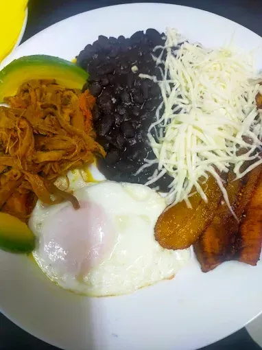 Un plato con carne deshebrada, frijoles negros, queso, huevo frito, aguacate y plátanos.