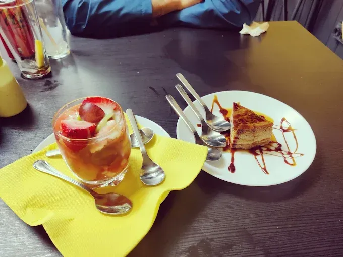 Postre: cóctel de frutas en vaso, rebanada de pastel en un plato con cucharas, sobre una mesa con una persona.