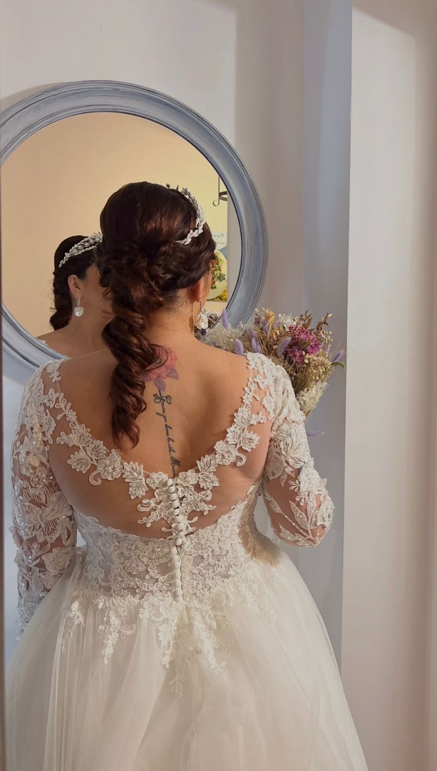 Novia con vestido de novia de encaje blanco, mirándose al espejo. Sosteniendo el ramo, con el tatuaje visible en la espalda.