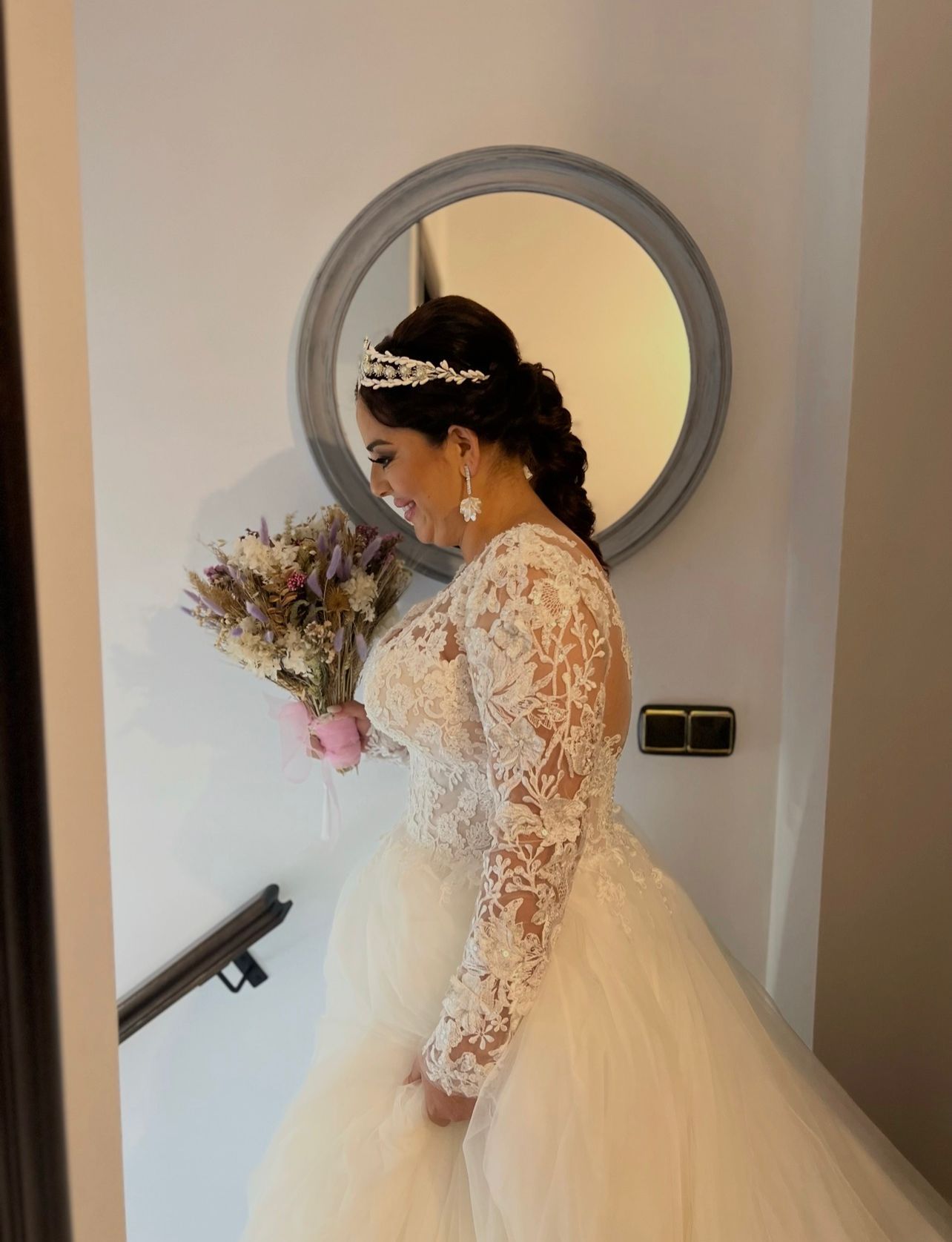 Novia con un vestido de novia de encaje blanco sosteniendo un ramo, de pie junto a una escalera, sonriendo.