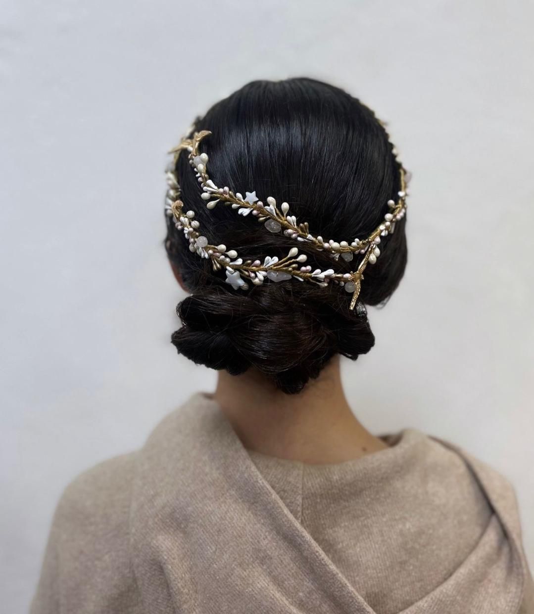 Mujer con cabello oscuro recogido en un moño, luciendo una diadema dorada con pequeños detalles florales blancos.