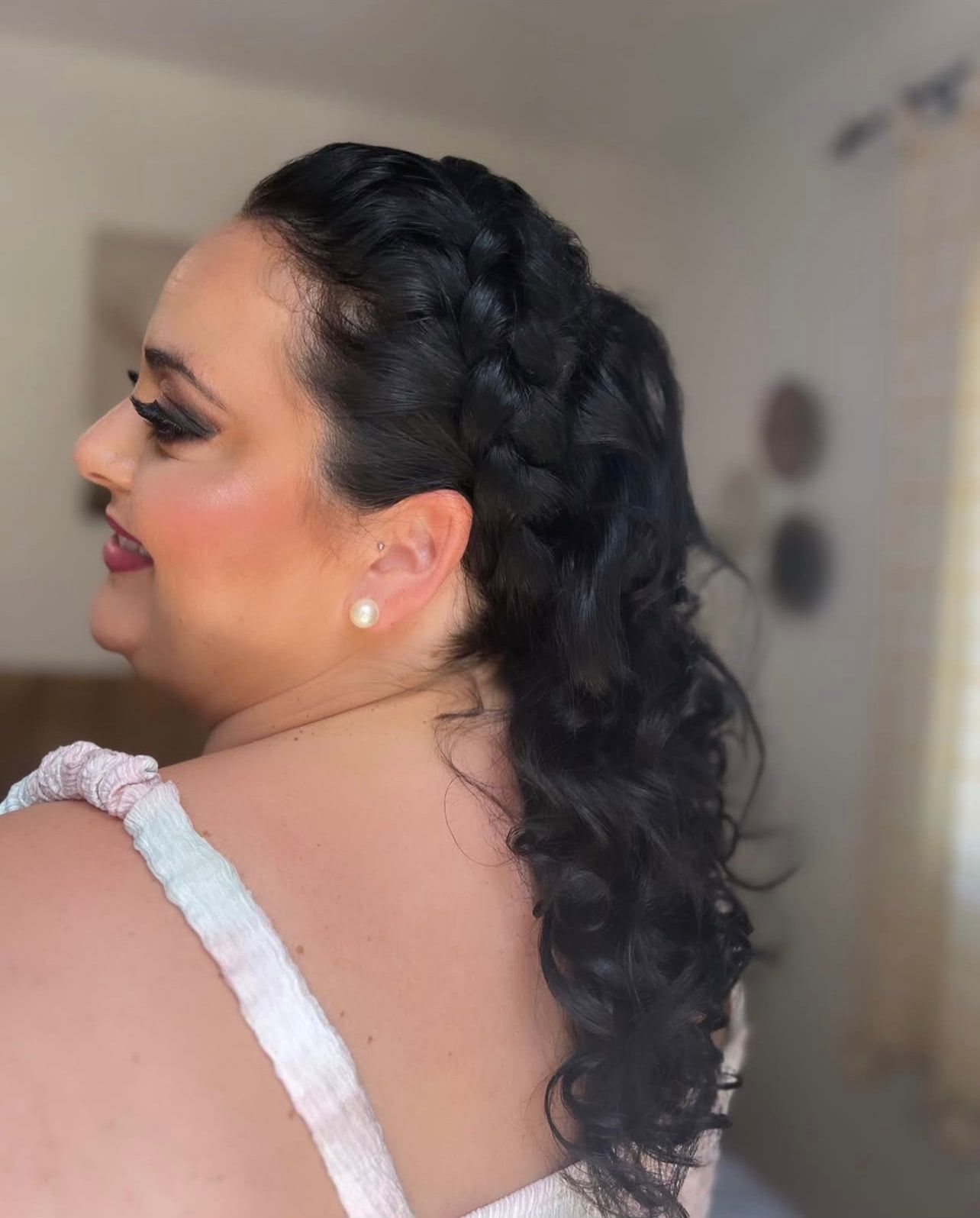 Mujer de cabello oscuro con un recogido trenzado. El cabello rizado le cuelga. Lleva maquillaje y vestido.