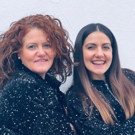 Dos mujeres sonríen, vistiendo blusas de lentejuelas negras contra una pared blanca.