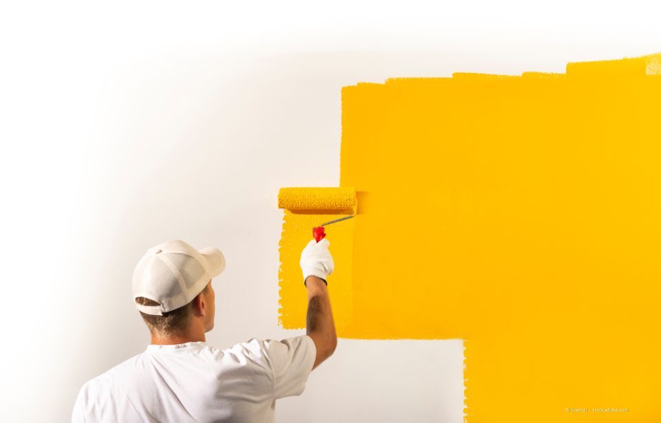 Ein Maler mit blauem Hut und Overall streicht eine weiße Wand mit einer Farbrolle. Leiter und Farbeimer stehen daneben.