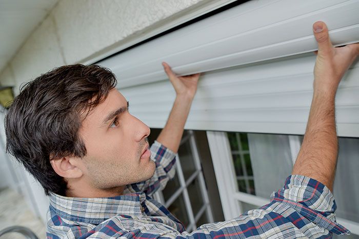 Un homme en chemise à carreaux installe des volets roulants blancs sur une fenêtre.