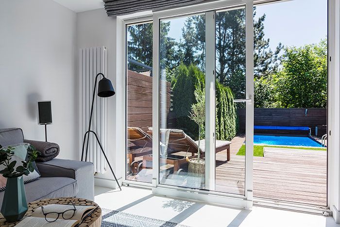 Des portes coulissantes en verre s'ouvrent sur une cour arrière avec piscine et mobilier de détente. L'intérieur est meublé d'un fauteuil gris et d'un lampadaire.
