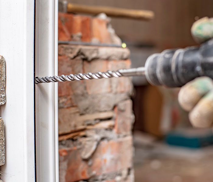 Une perceuse perce un cadre de porte blanc contre un mur de briques ; une main gantée tient la perceuse.
