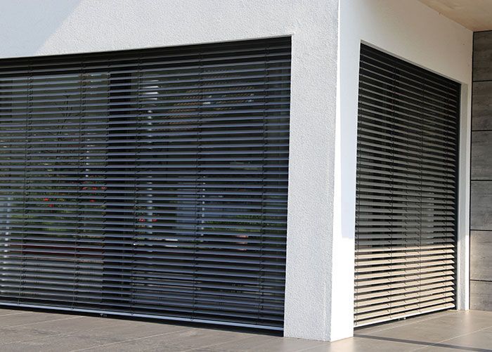 Vue extérieure d'un bâtiment aux fenêtres occultées par des volets horizontaux. La lumière du soleil se reflète sur les vitres.