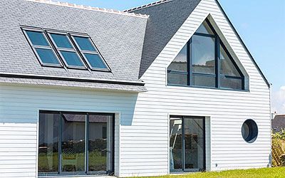 Maison blanche avec toit gris, grande fenêtre triangulaire, portes coulissantes en verre et puits de lumière.